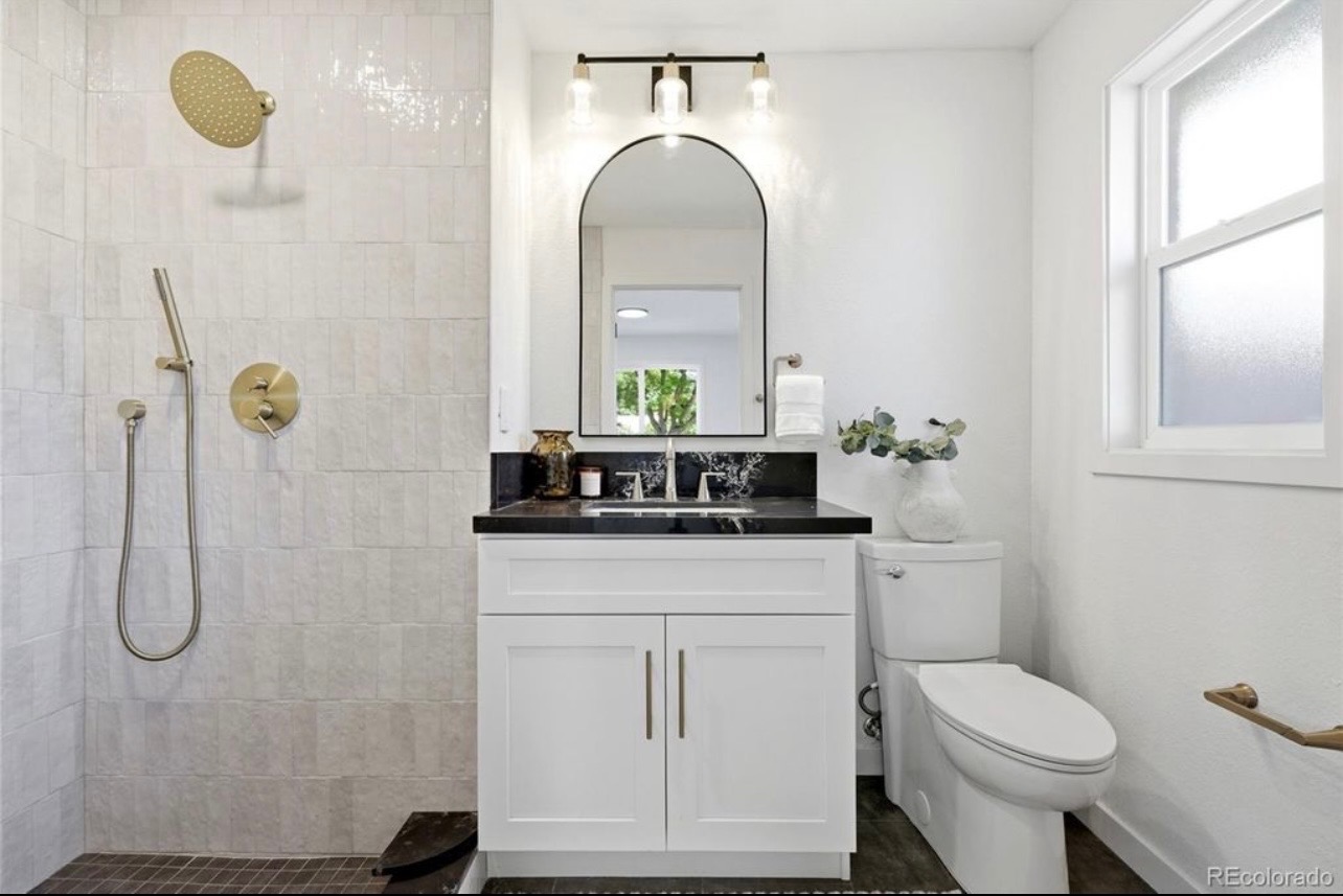 Zellige Shower with Gold Fixtures Walk-in shower with white zellige tile, brushed gold rain head and handheld, beside a white shaker vanity with black quartz counter and arched mirror